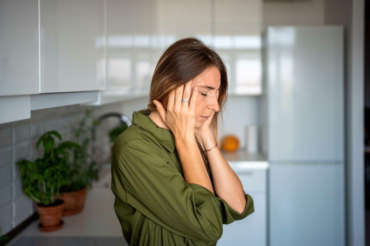 Eine Frau mit prämenstruellem Syndrom (PMS) lehnt an der Küchenzeile. Sie hält sich den Kopf, weil sie Kopfschmerzen hat und sich depressiv und ängstlich fühlt.